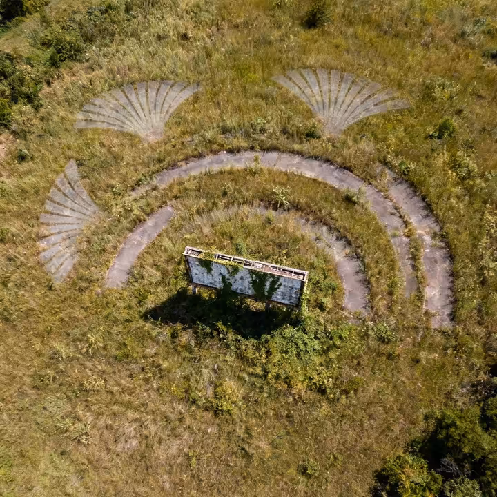 An aerial satellite view of an overgrown field showing the faint, curved outlines of former drive-in parking ramps.